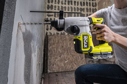 En person använda en grön och svart borrmaskin av märket RYOBI för att borra i en betongvägg.