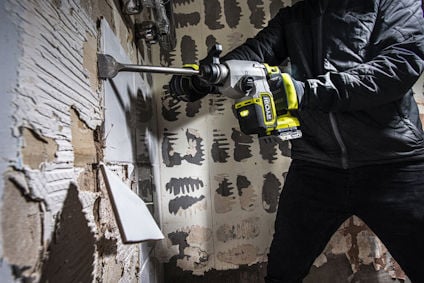 Person använder en batteridriven förstorningshammare på en vägg med en mejselbilaga, omgivning av riven puts och tegel.