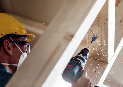 Person borrar i trä med en borrmaskin, bär skyddsglasögon och hjälm. Spån flyger runt.