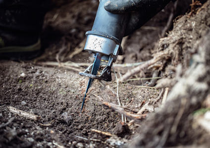 En hand i handske greppar ett blått verktyg med spets i kontakt med jorden, omgiven av rötter och jord.