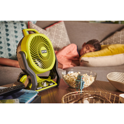 Grön RYOBI-fläkt på ett bord bredvid en skål med popcorn och en sovande person i bakgrunden.