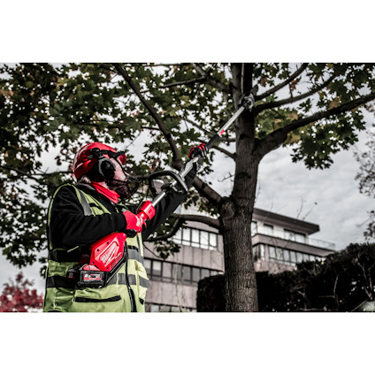 Person som beskär träd med en röd elektrisk grensåg, bär skyddsutrustning och väst, flerbostadshus i bakgrunden.