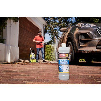 Flaska med skummedel från Ryobi på en uppfart, en person tvättar en bil i bakgrunden.