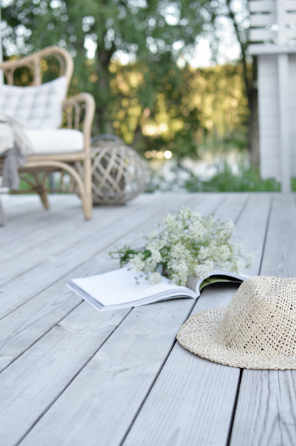 Stråhatt och en uppvikt bok på en träterrass med blommor i bakgrunden.