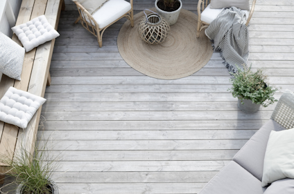 Utemöbler med dynor och kuddar på en träterrass, med växter och rottingdetaljer, sett snett uppifrån.