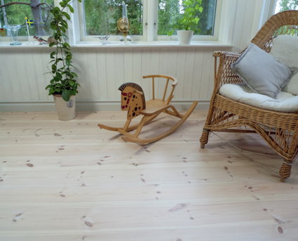 Gunghäst på ljust furugolv, grön växt i kruka och korgstol med kuddar framför ett fönster med oljelampa och prydnadssaker.