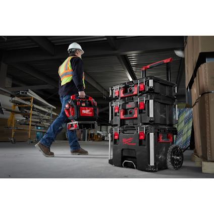 Man i arbetskläder och hjälm bär en verktygslåda, fler lådor på hjulförsedd vagn. Industrimiljö.