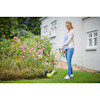 Kvinna i glasögon trimmar gräsmatta med grästrimmer bredvid en blomrabatt framför ett vitt hus.
