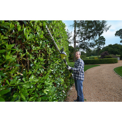 Person i rutig skjorta trimmar en hög häck med en batterdriven häcksax av märket RYOBI på en grusgång.