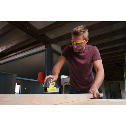 Person slipar träplanka med en eldriven slipmaskin i rum med träbjälkar. Bär skyddsglasögon.