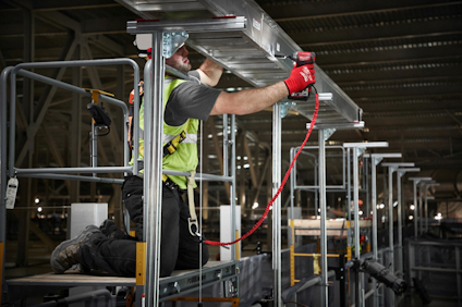 Person i arbetskläder monterar rör på ställning inomhus. Säkerhetssele och verktyg synliga.