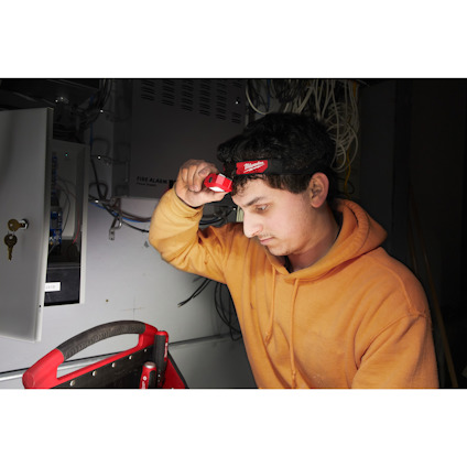Man in orange hoodie looks at electrical panel with a flashlight.