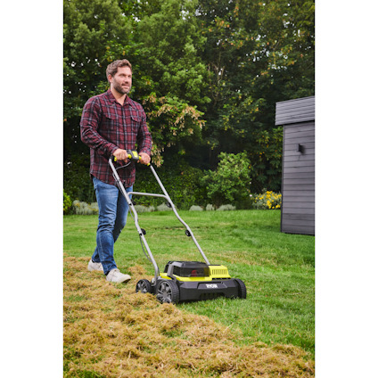En man i rutig skjorta och jeans går bakom en gräsklippare på en gräsmatta med träd i bakgrunden.