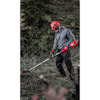 Person med skyddsutrustning använder en grästrimmer i en utomhusmiljö med gräs och buskar.