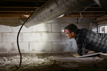 Person med pannlampa utforskar mörkt kryputrymme med exponerade rör och betongväggar.