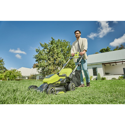 En person klipper gräset med en grön och svart gräsklippare på en solig dag, omgiven av hus och träd.