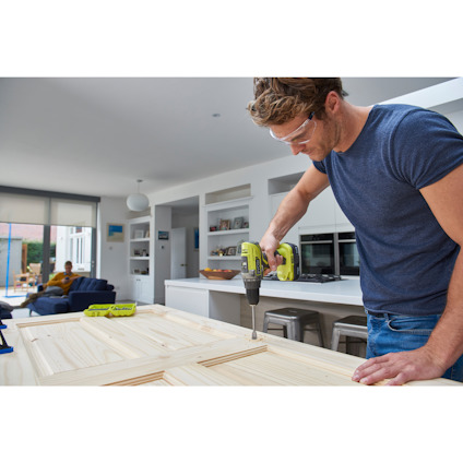 Person som borrar i en trädörr i ett modernt kök. En annan person syns i bakgrunden vid ett bord.