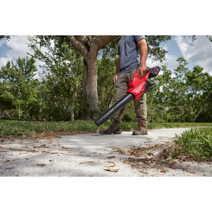 Person som använder en röd lövblåsare på en trottoar, med grönt gräs och träd i bakgrunden.