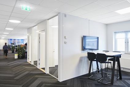 Kontorsmiljö med vita väggar, svarta möbler och en väggmonterad skärm. Flera dörrar längs korridoren.