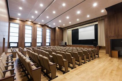 Stor föreläsningssal med träpaneler, radade stolar och projektorduk.