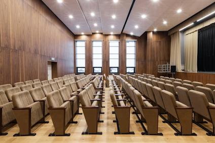 Stolar i rader i ett träklätt auditorium med fönster och scen i bakgrunden.