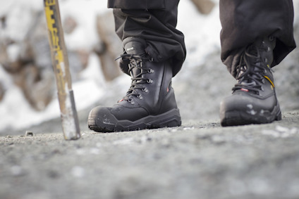 Två par svarta kängor med grov sula på grusig mark, angränsande till en stav.