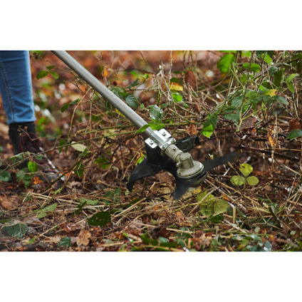 Person använder en gräs och slytrimmer med metallblad av märket Ryobi för att trimma buskar och grenar i skogsmiljö.