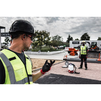 Person med svart hjälm och gul väst gestikulerar utomhus; bakgrund med fler arbetare och utrustning.