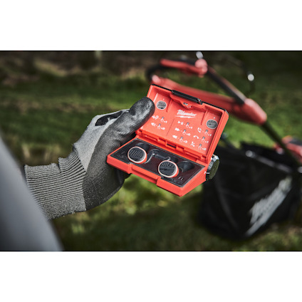 Person med handske håller ett rött Milwaukee-fodral med runda metallföremål i en trädgård.