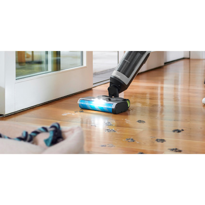 3-i-1 Skaftdammsugare av märket Ryobi rengör trägolv med djurspår på, bredvid glasdörrar.