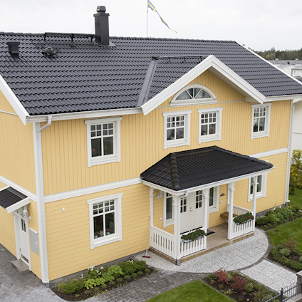 Gult trähus med svarta takpannor, vit dörr och vita fönsterkarmar. Stenlagd gång leder till entrén.