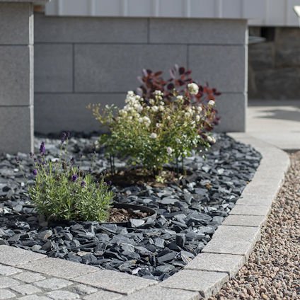 Blomrabatt med mörkt stenkross, gröna växter och lila blommor, kantad med stenar framför en mur.