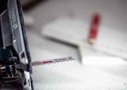 Sticksåg med ett Bosch-sågblad infogat, oskarp bakgrund med två vita plattor och en röd penna.