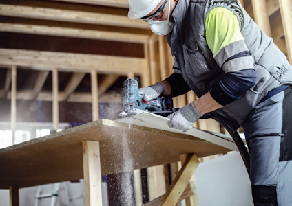 Person i skyddsutrustning använder elverktyg för att såga träskiva i byggnadsmiljö.