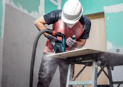 En person sågar en skiva med en eldriven såg på ett arbetsbord, iförd hjälm och skyddsmask.