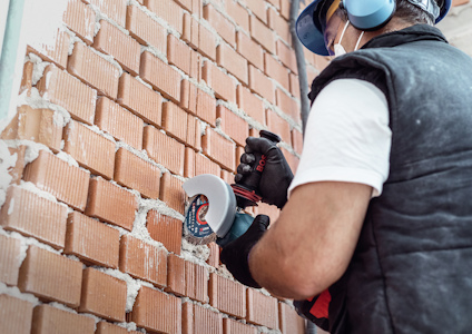 Person med hörselskydd och skyddsglasögon använder en vinkelslip mot en tegelvägg.