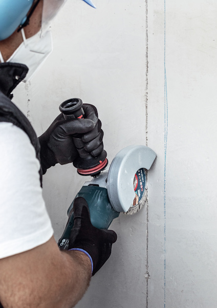 Person med skyddsglasögon och hjälm använder en väggfräs mot en vit yta.