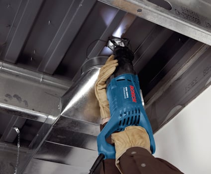 Person med handskar använder en blå elektrisk såg på en metallkanal i ett innertak.