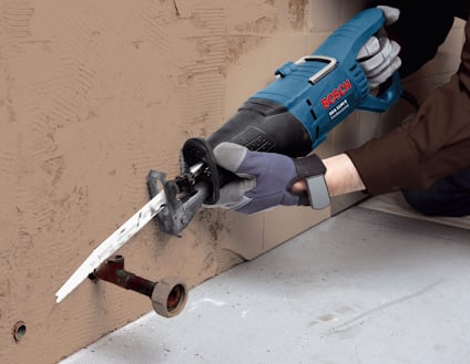 En person använder en blå elektrisk tigersåg för att skära ett rör genom en vägg.