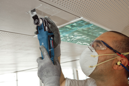 En person använder ett blått elverktyg för att arbeta på ett perforerat tak. Personen bär skyddsglasögon och ansiktsmask.