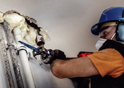 En person i hjälm och skyddsmask arbetar med rör som är täckta med isoleringsmaterial.