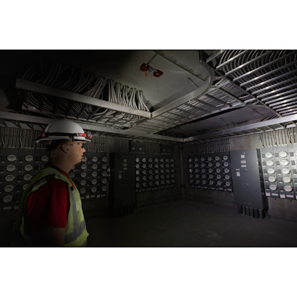 Person med hjälm och reflexväst står i rum fullt av mätare och kablar på taket.