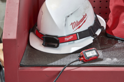 Vit hjälm med "Milwaukee" logotyp, fäst på rött stativ. Liten svart och röd enhet med kabel framför.