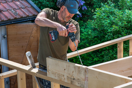 En person använder en borrmaskin på en träkonstruktion utomhus. Grönska syns i bakgrunden.