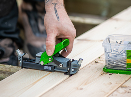 En person använder ett grönt och grått trallmonteringsverktyg för dolt montage och fäster träplankor. Skruvar ligger i en plastlåda bredvid.