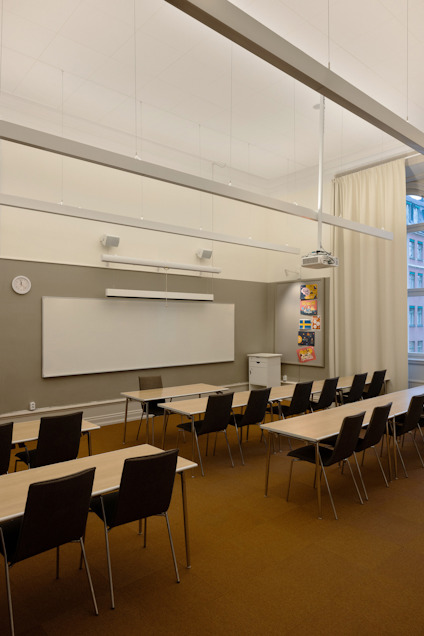 Klassrum med tomma skrivbord och stolar, vit skrivtavla, projektor och klocka på väggen. Fönster med utsikt mot byggnader.