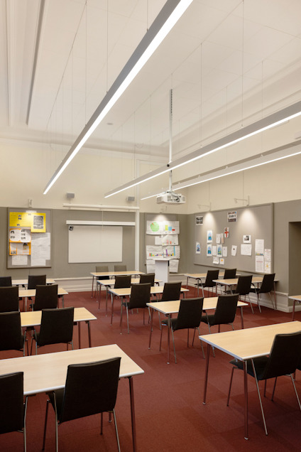 Klassrum med skrivbord och stolar, projektor i taket, anslagstavlor och väggdekorationer.