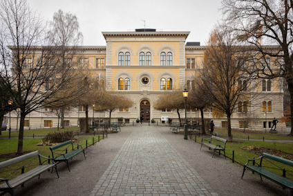 En trevåningsbyggnad med gul fasad, träd och bänkar längs en stenbelagd gångväg i förgrunden.