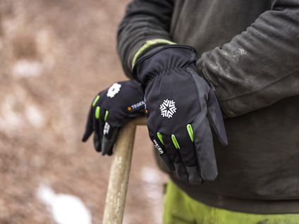 Person i mörk tröja som håller ett träverktyg, bär svarta handskar med gröna detaljer och vit logga.