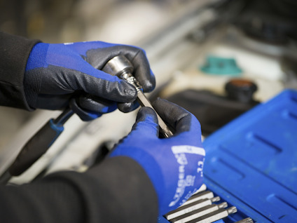 Händer i blåa handskar håller i ett verktyg nära en motor. Verktygslåda syns i bakgrunden.